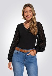 Schwarzes Oberteil mit floralen Spitzendetails an den langen Ärmeln, V-Ausschnitt, kombiniert mit blauen Jeans und einem braunen Gürtel, der eine glatte Textur und eine figurbetonte Passform aufweist.