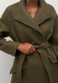 Osoba zapínající pásek olivově zeleného vlněného kabátu s širokými klopami a klopovými kapsami.