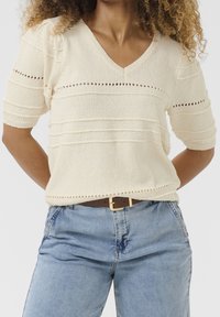 Žena s kadeřavými vlasy má na sobě krémový pletený svetr s výstřihem do V a krátkým rukávem, k tomu světle modré džíny s vysokým pasem a hnědym páskem.