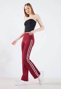 Pantalons bordeaux taille haute avec des rayures blanches sur les côtés, fabriqués en tissu extensible. Associés à un haut noir moulant sans manches et des baskets blanches.