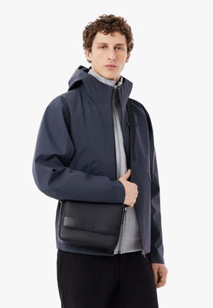 Joven con chaqueta oscura con capucha sobre una sudadera gris con cremallera, sosteniendo una bolsa cruzada negra con textura y solapa.