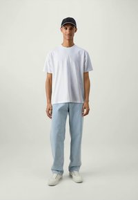 Mladý muž v černé kšiltovce, bílém tričku, světle modrých džínách a bílých teniskách stojící proti jednolitému světlému pozadí.