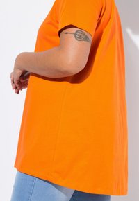 Orange kortærmet t-shirt lavet af bomuld, med en afslappet pasform og en lige kant. Båret med blå jeans, der viser en glat tekstur.