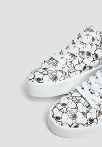 Baskets blanches avec des visages de cartoon Snoopy en noir et blanc imprimés partout, dotées de lacets blancs et de semelles épaisses blanches.