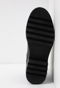 Semelle de chaussure noire avec surface texturée, motifs nervurés et talon arrondi. Le côté présente une tige lisse avec un design minimaliste. Taille 37 visible.