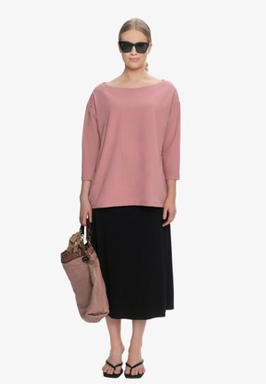 Kobieta ubrana w oversize różowy top z długim rękawem, czarną spódnicę midi, czarne sandały, ciemne okulary przeciwsłoneczne i trzymająca dużą różową torebkę.