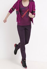 Fialový sportovní outfit skládající se z bundy na zip s černými a oranžovými akcenty, odpovídajícího trička s logem a přiléhavých kalhot, doplněný černými teniskami.