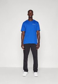 Vīrietis stāv zilanā Champion T-kreklā, melnās biksēs un baltos sporta apavus pret vienkāršu gaišu fona.