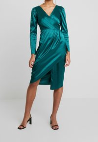 Smaragdgrünes Satin-Wickelkleid mit langen Ärmeln, V-Ausschnitt, Leopardenmuster und einem seitlichen Schlitz. Kombiniert mit schwarzen High Heels.