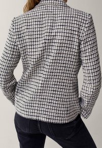 Femme portant un blazer cintré à carreaux noir et blanc à manches longues et un pantalon noir, vue de dos.