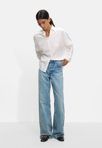 White long-sleeve button-up shirt, loose fit. Light blue wide-leg jeans, faded texture. Black shoes with studded accents.