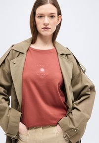 Γυναίκα που φοράει πουκάμισο χρώματος σκουριάς με το κείμενο «Amar el sol» και γραφικό ήλιου, με στρώσεις από γκρι τραντς και μπεζ παντελόνι, τα χέρια στις τσέπες.