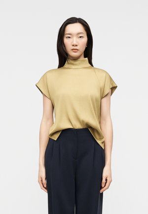 Žena v béžovém krátkém topu s vysokým límcem, zastrčeném do černých širokých kalhot s vysokým pasem, stojící před jednobarevným pozadím.