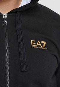 Svart zip-up hoodie i bomull med en vävd dragsko. Har en guldig EA7 Emporio Armani-logotyp på vänster bröst.