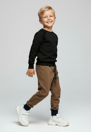Uśmiechnięty młody chłopiec idący, ubrany w czarną bluzę, brązowe spodnie dresowe, czarne skarpetki i białe trampki na jednolitym szarym tle.