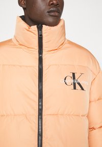 Giacca puffer arancione chiaro con colletto alto, zip nera e grande logo "Calvin Klein Jeans" in nero sul petto.