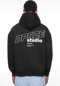 Sort hættebluse med stor hvid tekst på bagsiden, der står "DPSZE studio" samt ekstra sætninger nedenunder. Fremstillet af blødt stof med en kængurulomme.