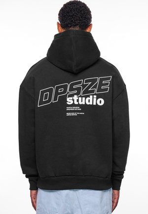 Sort hættebluse med stor hvid tekst på bagsiden, der står "DPSZE studio" samt ekstra sætninger nedenunder. Fremstillet af blødt stof med en kængurulomme.