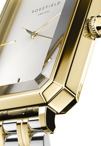 Montre en or et argent avec un boîtier rectangulaire. Cadran argenté texturé, aiguilles dorées et couronne détaillée. Fini brillant, éléments de design moderne.