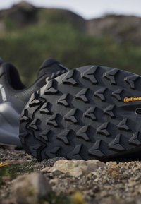 Gros plan de la semelle extérieure noire d'une chaussure de randonnée avec un motif de bande de roulement texturé reposant sur un sol rocheux en plein air.