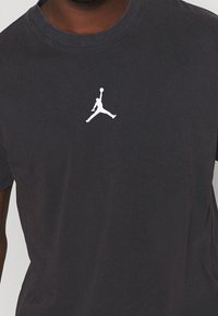 Person som bär en svart t-shirt med en liten vit Jumpman-logotyp centrerad på bröstet, visar endast överkroppen.