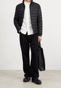 Osoba v černé bundě s péřovou izolací, bílé košili s knoflíky, černých kalhotách, černých botách, držící černou koženou tašku po svém boku.