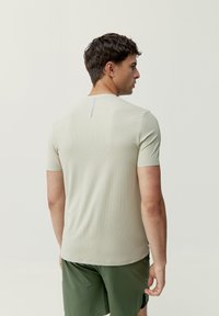 Īss piedurkņu, gaiši bēšs sporta krekls ar teksturētu audumu. Iezīmējas ar smalku vertikālu šuvi aizmugurē un pieguļošu dizainu.