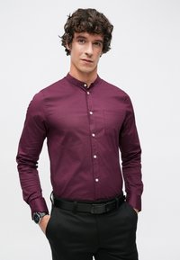 Bordowa koszula zapinana na guziki z kołnierzem typu band, pojedynczą kieszenią na piersi i długimi rękawami. W zestawie z czarnymi spodniami i czarnym paskiem.