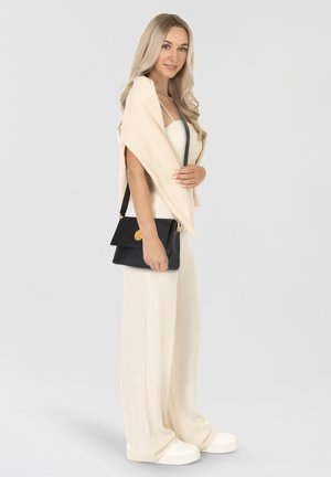 Beige knitted shawl drapes over a light cream jumpsuit. Model carries a black leather crossbody bag with a gold clasp. White sneakers complete the look.