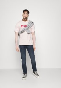Svijetlo ružičasta Levi's majica s crvenim logotipom, sivi dukserica prebačena preko ramena, plave traperice i crne tenisice s bijelim potplatima vidljive.