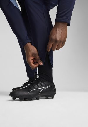 Botas de futebol pretas com parte superior texturizada e detalhes brancos, combinadas com calças de treino azul-marinho; uma mão ajusta o fecho no tornozelo.
