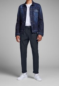 Jeansjacka med mörk tvätt, vit t-shirt med rund hals, texturerade mörka byxor och vita sneakers. Modellen står mot en neutral bakgrund.