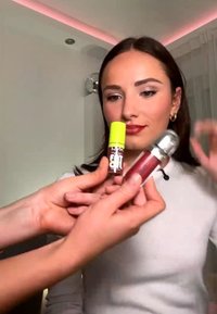 Femme aux cheveux foncés et au haut blanc observe deux gloss à lèvres, un avec un bouchon jaune et un avec un bouchon argenté, tenus près de son visage.
