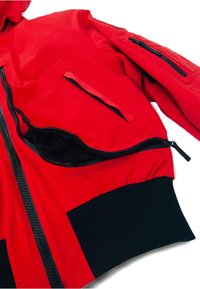 Piros dzseki fekete részletekkel, elején cipzárral, több cipzáras zsebbel az ujjakon, és bordázott mandzsettákkal a kényelmes illeszkedésért.