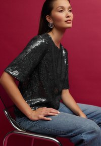 Sort perlesyet top med korte ærmer, har en tekstureret overflade, parret med lyseblå denimjeans og en metallisk stol, sat mod en pink baggrund.