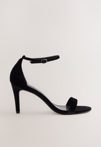 Crna baršunasta sandala na visoku petu s tankim remenom oko članka i okruglim metalnim detaljem. Odlikuje je minimalistički dizajn i otvoreni prednji dio.
