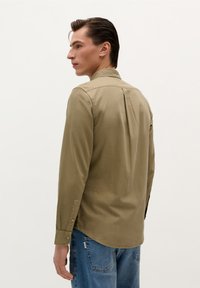 Camicia khaki a maniche lunghe con colletto classico, polsini con bottone e vestibilità rilassata. Realizzata in un tessuto liscio con una leggera lucentezza.