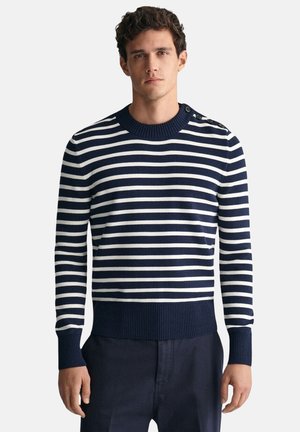 Mann trägt marineblauen und weißen horizontal gestreiften Pullover mit Knöpfen auf der linken Schulter und marineblaue Hose, steht vor einfachem Hintergrund.