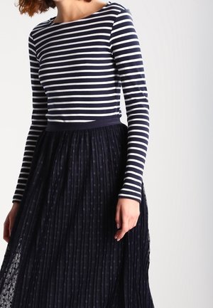 Femme portant un haut ajusté à manches longues à rayures horizontales bleu marine et blanches, associé à une jupe plissée taille haute bleu marine avec des pois subtils.
