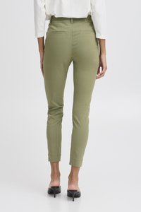 Femme portant un pantalon slim vert olive et des mules à talons hauts noires, debout de dos sur un fond blanc uni.