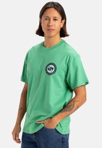 Camiseta de algodón verde con un parche de logo redondo en el pecho. De manga corta, corte relajado y cuello redondo, combinada con jeans azules.