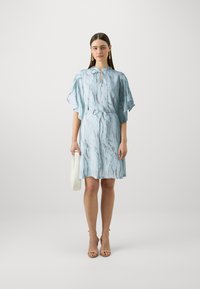 Robe bleu clair avec des manches fluides, un décolleté en gouttière et une taille élastique. Présente un imprimé marbré et est associée à un sac à main blanc.