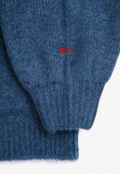 Manche de pull bleu tricoté avec des lettres rouges brodées "BC" près du poignet, montrée pliée sur un fond blanc.