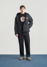 Μαύρο μπουφάν με φερμουάρ και υφασμένο κουκούλι, φορεμένο πάνω από μαύρο μπλουζάκι με κυκλικό σχέδιο. Συνδυασμένο με μαύρο παντελόνι και μπεζ παπούτσια.