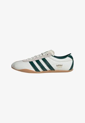 Zapatilla Adidas de perfil bajo blanca con rayas verde oscuro, suela de goma, zona de cordones en ante y la marca "adidas Tokyo" en el lateral.