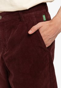 Burgundy corduroy pants with vertical ribbed texture, featuring a side pocket and a green brand tag. Metal button closure at the waist.