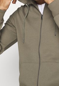 Sudadera con capucha y cremallera en verde oliva, hecha de tela suave. Cuenta con capucha con cordón, cremallera frontal y bolsillos tipo canguro. Textura lisa con puños acanalados.