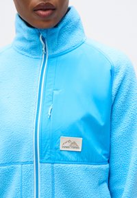 Μπλε fleece μπουφάν με φερμουάρ και ψηλό γιακά, διαθέτει υφή υφάσματος και λεία επάνω περιοχή. Περιλαμβάνει λογότυπο στο στήθος.