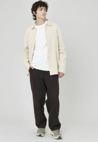 Chemise beige à boutons avec un tissu texturé, t-shirt blanc en dessous, et pantalon foncé. Le mannequin porte des baskets, debout avec une main dans la poche.