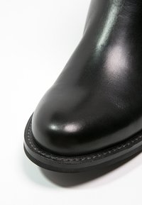 Gros plan d'une botte en cuir noir avec un bout arrondi et une semelle épaisse cousue sur fond blanc.